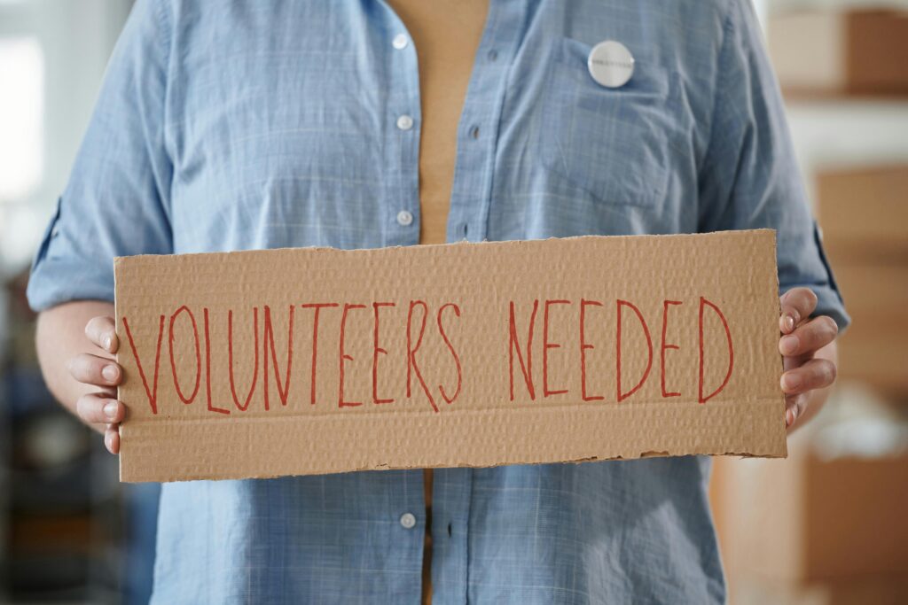 pexels-photo-6994992-6994992 Person holding a sign reading 'Volunteers Needed' to encourage community support.